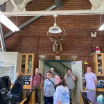 A group of college students stand in a lab. There is a large deer head mounted on the wall. On the black-topped lab tables are covered microscopes.